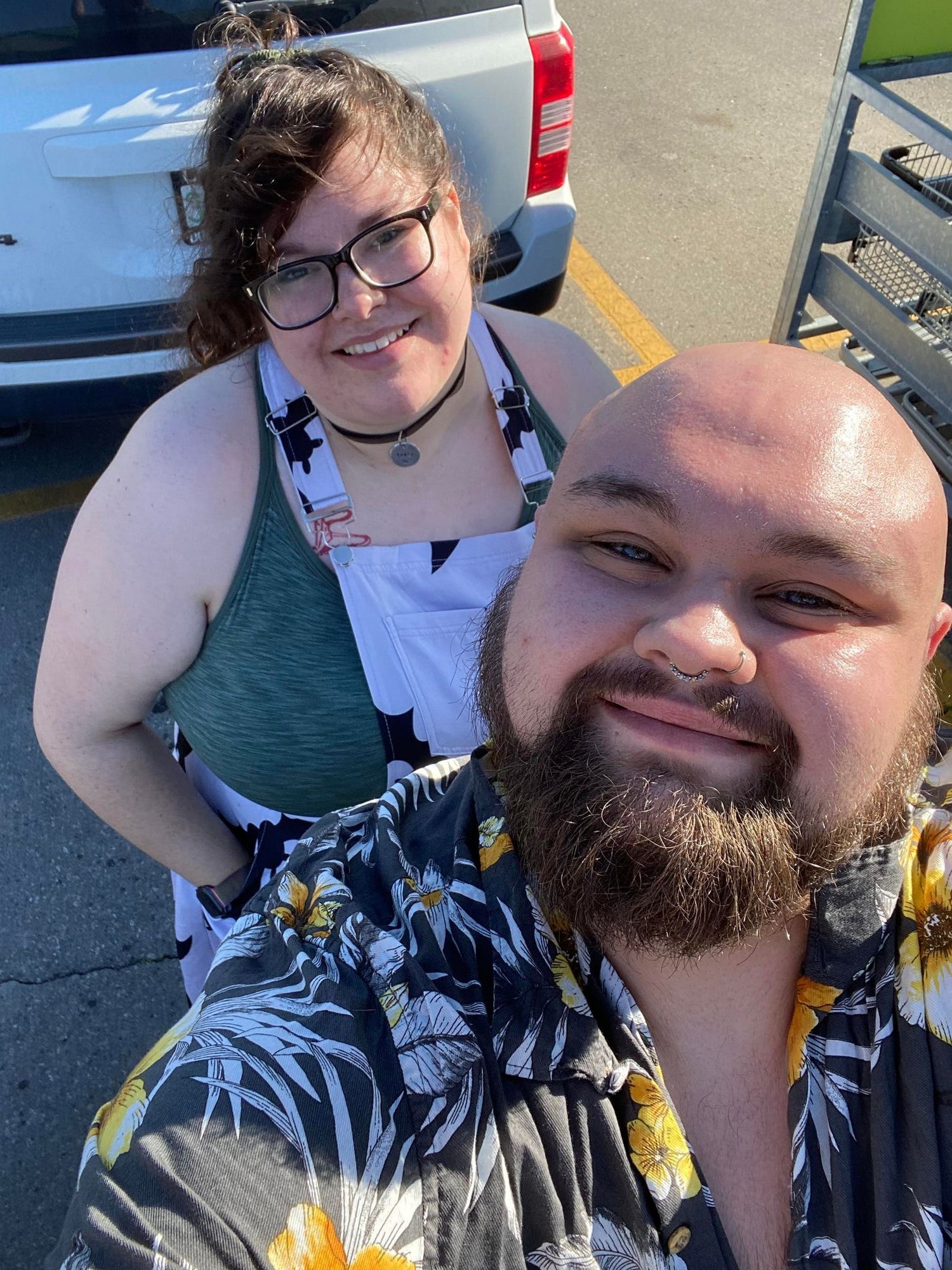 a selfie of two people. standing in the back is a person with long brown hair and light skin. they are wearing glasses and smiling at the camera. in the front is a bald, bearded white person with a septum piercing. they are smiling up at the camera.